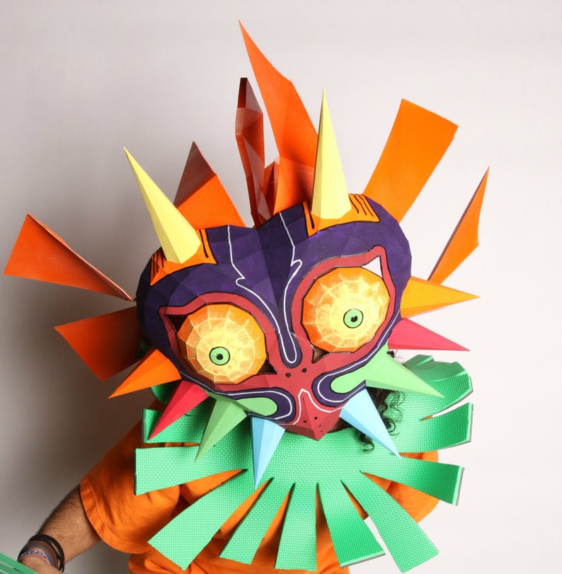 May include: A papercraft mask of a character with a large, colorful, and detailed design. The mask has a purple, red, and green face with yellow eyes and sharp teeth. The mask is surrounded by orange and yellow spikes.
