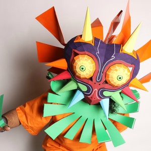 May include: A person wearing a paper mache mask of Majora's Mask from the Legend of Zelda video game series. The mask is colorful and detailed, with sharp points and a menacing expression. The person is wearing an orange shirt and green paper mache gloves.