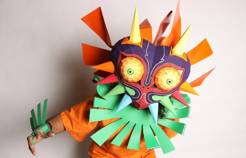 May include: A person wearing a paper mache mask of Majora's Mask from the Legend of Zelda video game series. The mask is colorful and detailed, with sharp points and a menacing expression. The person is wearing an orange shirt and green paper mache gloves.