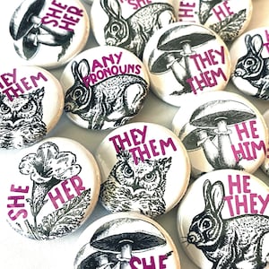 Peut inclure: Un ensemble de boutons ronds avec différentes illustrations et textes. Les boutons présentent des images de hiboux, de lapins, de champignons et de fleurs. Le texte sur les boutons comprend "They/Them", "She/Her", "He/Him", "Any Pronouns", et "They She".