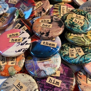 May include: A collection of colourful round buttons with various images and text. The text on the buttons includes "she her", "they them", "he him", and "he him".