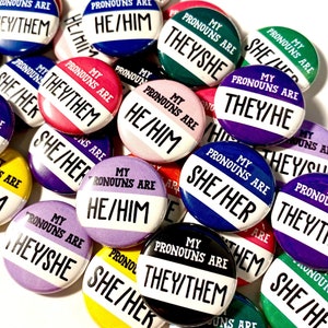 May include: A collection of colourful button badges with the text "My Pronouns Are" followed by different pronoun sets, including they/them, he/him, she/her, they/she, and they/he.
