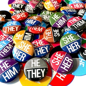 May include: A collection of colourful round buttons with different pronoun combinations, including "She/Her", "He/Him", "They/Them", and "She/They".