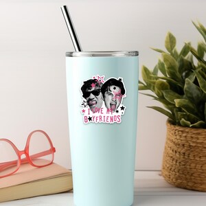 Op de afbeelding: Een lichtblauwe roestvrijstalen beker met een metalen rietje, voorzien van een sticker van twee gezichten met zonnebrillen en uitgestoken tongen, omringd door roze en zwarte sterren en de tekst I LOVE MY BOYFRIENDS in roze en zwarte letters.