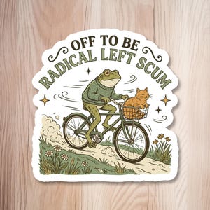May include: A die-cut sticker featuring a green frog wearing a green jumper, riding a bicycle on a dirt path. An orange cat sits in the front basket. The text "OFF TO BE RADICAL LEFT SCUM" is arched above the frog, surrounded by small stars.