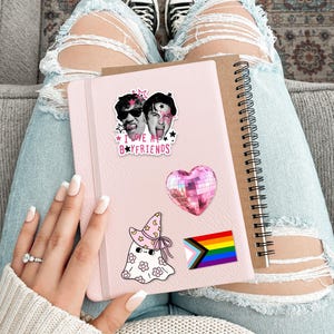 Op de afbeelding: Een lichtroze notitieboek versierd met vier stickers: een zwart-witfoto van twee personen met de tekst I LOVE MY BOYFRIENDS, een roze hartvormige discobal, een witte geest met een roze heksenhoed met bloemen en een regenboogvlag.