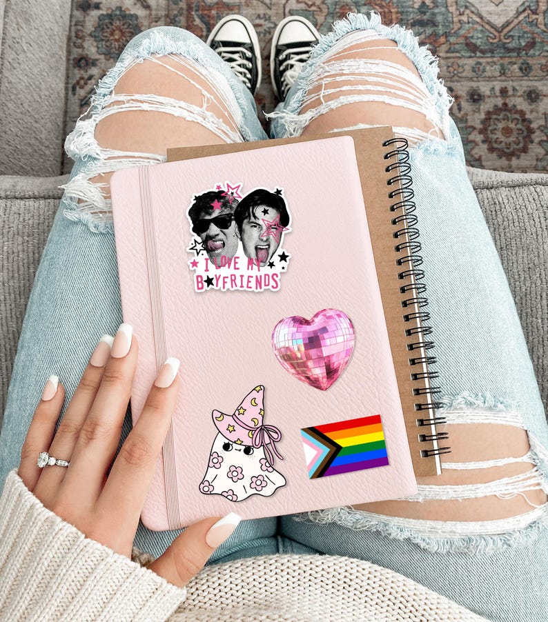Op de afbeelding: Een lichtroze notitieboek versierd met vier stickers: een zwart-witfoto van twee personen met de tekst I LOVE MY BOYFRIENDS, een roze hartvormige discobal, een witte geest met een roze heksenhoed met bloemen en een regenboogvlag.