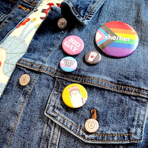 May include: A denim jacket with a variety of buttons pinned to it. The buttons feature different designs, including a rainbow flag with the text "she/her", a pink button with the text "BIG GAY", a blue button with the text "PROTECT TRANS KIDS", a brown button with a coffee cup design, and a yellow button with a llama design.