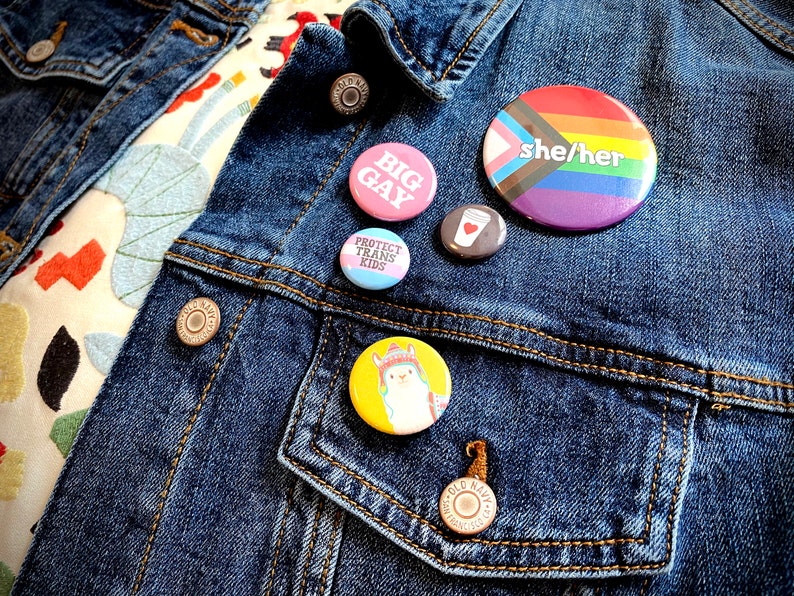 May include: A denim jacket with a variety of buttons pinned to it. The buttons feature different designs, including a rainbow flag with the text "she/her", a pink button with the text "BIG GAY", a blue button with the text "PROTECT TRANS KIDS", a brown button with a coffee cup design, and a yellow button with a llama design.