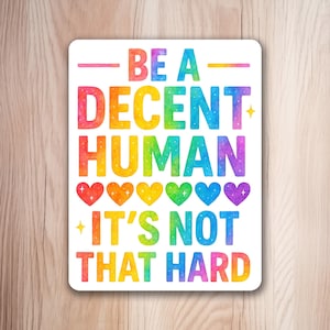 May include: A rectangular sticker with rounded corners featuring the text BE A DECENT HUMAN IT'S NOT THAT HARD in a rainbow-coloured watercolour gradient. The design includes six small hearts in rainbow colours and four small star icons.