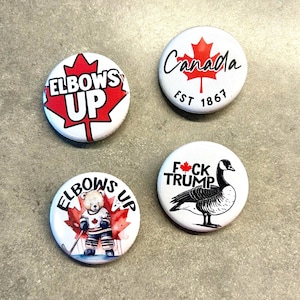 May include: Four round white button badges with different designs. The first badge has a red maple leaf with the text "Elbows Up". The second badge has a red maple leaf with the text "Canada Est 1867". The third badge has a red maple leaf with the text "Elbows Up" and a cartoon bear wearing a hockey uniform. The fourth badge has a red maple leaf with the text "F*ck Trump" and a cartoon goose.