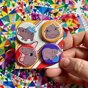May include: Set of four round button badges featuring cartoon hippos in various poses. The badges are colourful and feature a white background with a red, orange, purple, and blue border. The hippos are depicted in a playful and whimsical style.