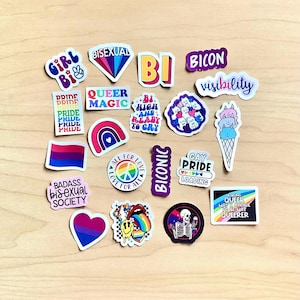 May include: A selection of vibrant stickers with diverse designs and text. The stickers display phrases such as "Pride," "Queer Magic," and "Bisexual." Other designs include a rainbow, a heart, and a skeleton holding a drink. The stickers are on a light brown surface.