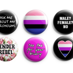 May include: Six circular pin-back badges. Top row: black badge with white text 'ASK ME ABOUT MY PRONOUNS', a badge with horizontal pink, white, purple, black, and blue stripes, and a black badge with white text 'MALE? FEMALE? NO'. Bottom row: floral badge with black text 'GENDER REBEL', a pink badge with black text 'too fab for your binary', and a black badge with a heart featuring pink, white, purple, black, and blue stripes.