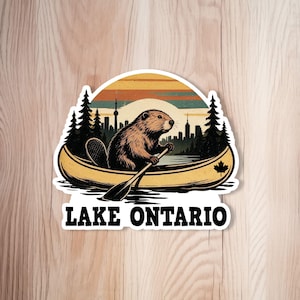 Op de afbeelding: Een gestanste sticker met een bruine bever die in een gele kano op het water peddelt. De achtergrond toont een stadssilhouet, dennenbomen en een zonsondergang met horizontale strepen. Een esdoornblad staat op de kano. De tekst luidt LAKE ONTARIO.