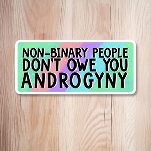 Może przedstawiać: Prostokątna naklejka z napisem "NON-BINARY PEOPLE DON'T OWE YOU ANDROGYNY" pogrubioną czarną czcionką. Tło to mieszanka odcieni turkusu, fioletu i różu. Naklejka ma białą obwódkę.