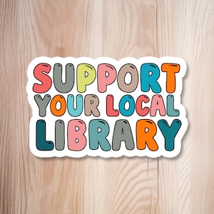 May include: A colourful sticker with the text "SUPPORT YOUR LOCAL LIBRARY" in bubble letters. The text is outlined in white and features a variety of colours, including pink, orange, blue, and green. The sticker is on a wooden background.