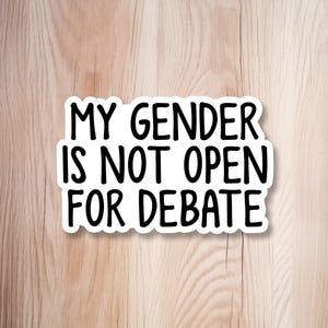 May include: A white rectangular sticker with rounded corners featuring the black text MY GENDER IS NOT OPEN FOR DEBATE in a bold, sans-serif font. The sticker has a clean white border and is presented as a flat graphic element on a light background.