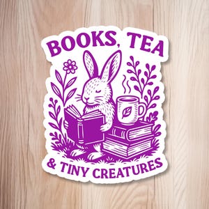 May include: A purple sticker featuring a rabbit reading a book, with a cup of tea and the text "BOOKS, TEA & TINY CREATURES." The design includes floral elements and a stack of books, creating a whimsical, book-themed aesthetic.