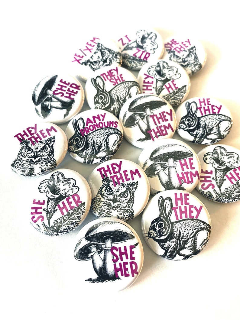 May include: A collection of round buttons with various illustrations and text. The buttons feature images of owls, rabbits, mushrooms, and flowers. The text on the buttons includes "They/Them", "She/Her", "He/Him", "Any Pronouns", and "They She".