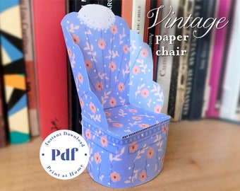 May include: A printable paper chair model with a vintage floral design. The chair is light blue with pink flowers and leaves. The text "Vintage paper chair" is written in cursive above the chair. The text "Instant Download Pdf Print at Home" is written in a circle below the chair.