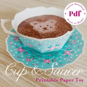 Puede incluir: Un juego de taza y platillo de papel con un diseño floral turquesa. La taza está llena de una sustancia marrón con un diseño de cara de oso. La imagen incluye el texto "Descarga instantánea PDF Imprimir en casa" y "Taza y platillo Juguete de papel imprimible."