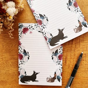 May include: Two lined notepads with floral borders and illustrations of a black fox and a brown cat. The floral design includes burgundy and blue-green leaves and flowers. A black pen rests on a wooden surface.
