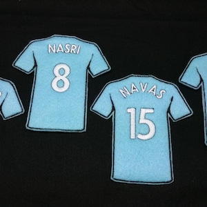 May include: Four light blue jersey-shaped patches featuring white embroidered names and numbers. The patches display the names AGÜERO with number 16, NASRI with number 8, NAVAS with number 15, and TOURÉ with number 42, all outlined in black thread.
