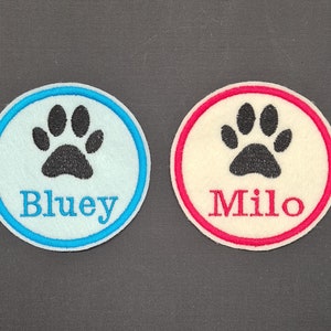 May include: Two circular patches with embroidered paw prints. One is light blue with a dark blue border and the name "Bluey" in blue. The other is off-white with a red border and the name "Milo" in red.