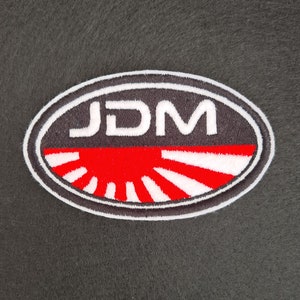 May include: A white oval patch with the word "JDM" in white letters on a black background. The patch has a red and white rising sun design in the center.