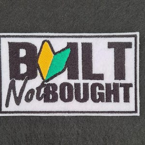 May include: White embroidered patch with black text that reads "BUILT Not BOUGHT". A green and yellow stylized logo is to the left of the text.
