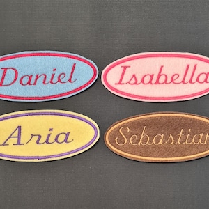 May include: Four oval-shaped fabric patches with embroidered names. The patches are blue with red stitching, pink with red stitching, yellow with purple stitching, and brown with yellow stitching. The names are "Daniel", "Isabella", "Aria", and "Sebastian".