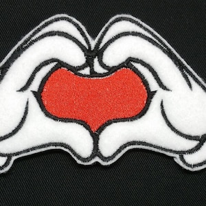 May include: White felt hands forming a heart shape with a red felt heart inside. The hands are outlined in black thread.