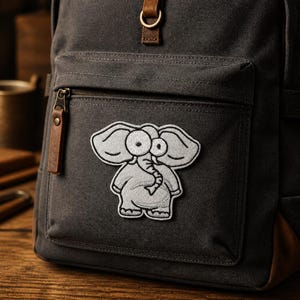 May include: A dark gray canvas backpack featuring a light gray embroidered patch of a cartoon elephant with large eyes and a trunk. The backpack includes a front zippered pocket with a brown leather pull tab and a brown leather strap detail above.