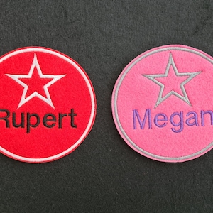 May include: Two round felt badges with white stars and embroidered names. The red badge says "Rupert" and the pink badge says "Megan".