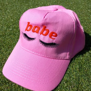 May include: Pink baseball cap with the word "babe" embroidered in orange with black eyelashes above the word.