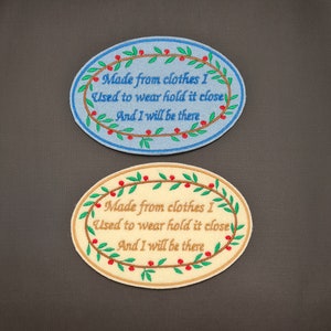 May include: Two oval-shaped embroidered patches with a green leafy wreath and red berries. The text on the patches reads "Made from clothes I Used to wear hold it close And I will be there". The top patch is on a light blue background and the bottom patch is on a cream background.