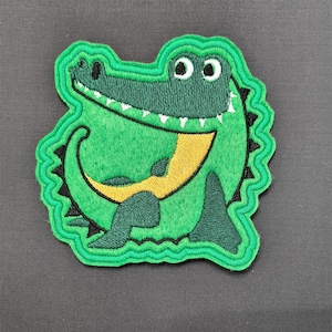 May include: Green felt applique of a smiling alligator with yellow teeth and a yellow belly. The alligator is outlined in green thread.