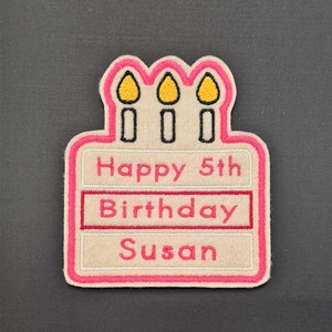May include: A pink and white birthday cake patch with three yellow candles. The cake has the text "Happy 5th Birthday Susan" on it.