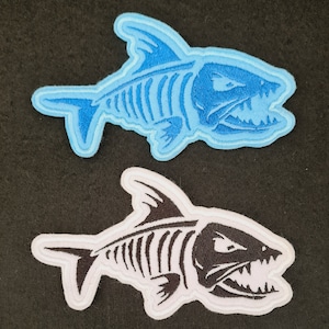 May include: Two embroidered iron-on patches of a skeleton fish with an open mouth. One patch is blue and the other is black.