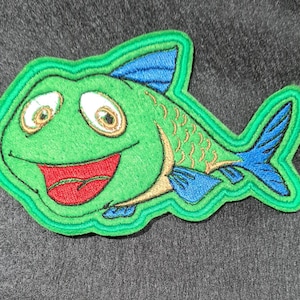 May include: Green embroidered fish patch with a smiling face, blue fins, and gold scales. The patch has a green outline.