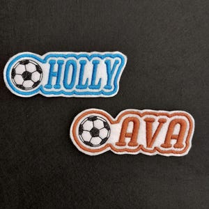 May include: Two embroidered patches featuring soccer ball designs. One patch reads "HOLLY" in blue lettering with a white outline, and the other reads "AVA" in orange lettering with a white outline. Both patches include a black and white soccer ball.