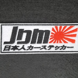 May include: White embroidered patch with black text that reads "JDM" and "日本人カー ステッカー" which translates to "Japanese Car Sticker". The patch also features a red and white rising sun design.