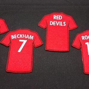 May include: Four red fabric patches shaped like sports jerseys. Each patch features white embroidered text and numbers. The patches display the names V. PERSIE with 20, BECKHAM with 7, RED DEVILS, and ROONEY with 10, all arranged on a dark surface.