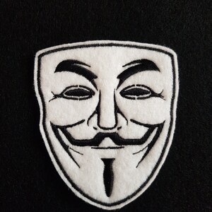 May include: A white felt patch with a black embroidered image of the iconic Guy Fawkes mask. The mask features a wide grin, a mustache, and a goatee.