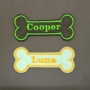 May include: Two dog bone shaped patches with embroidered names. The top patch is black with a green outline and the name "Cooper" embroidered in green. The bottom patch is light blue with an orange outline and the name "Luna" embroidered in orange.
