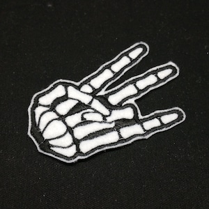 May include: White embroidered patch of a skeleton hand giving the peace sign, on a black background.
