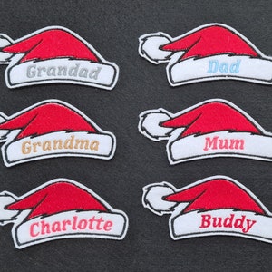 May include: Six red felt Christmas hat patches with white trim. The patches are embroidered with the names "Granddad", "Dad", "Grandma", "Mum", "Charlotte", and "Buddy".
