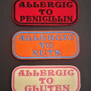 May include: Three rectangular fabric patches with black stitching around the edges. The top patch is red with black text that reads "ALLERGIC TO PENICILLIN". The middle patch is orange with blue stitching around the edges and black text that reads "ALLERGIC TO NUTS". The bottom patch is beige with pink stitching around the edges and black text that reads "ALLERGIC TO GLUTEN".