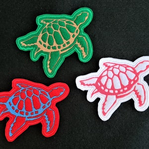 May include: Three felt sea turtle patches in different colours. A green turtle, a red turtle, and a white turtle with pink outlines.
